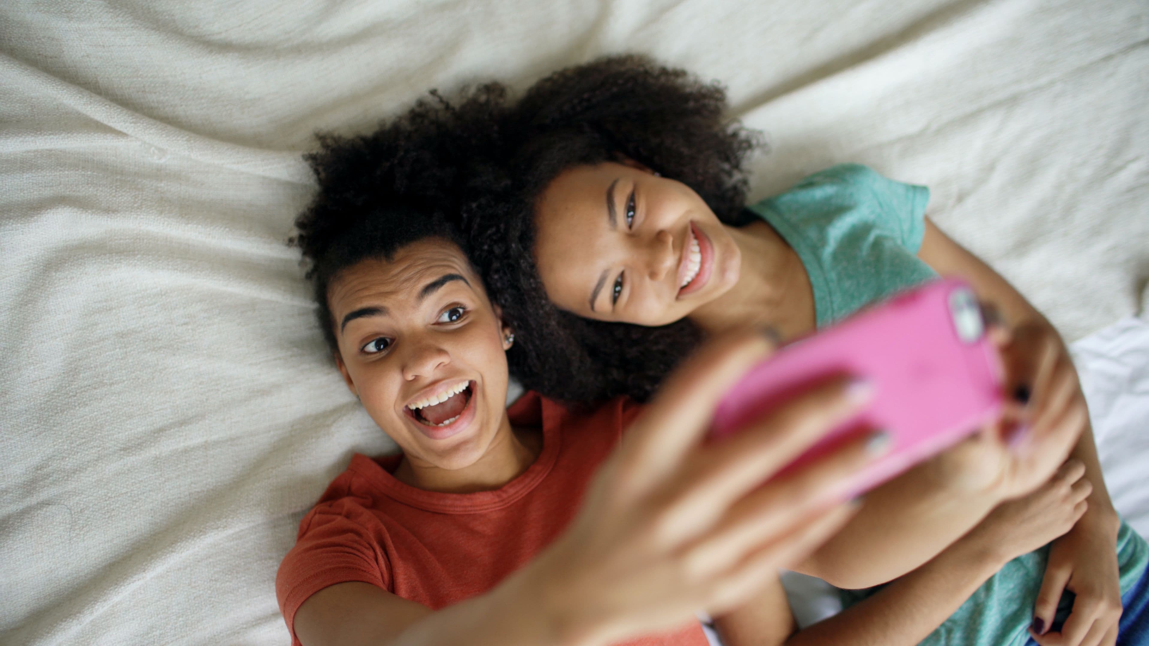 Two smiling teenage girls lying on a bed taking a selfie