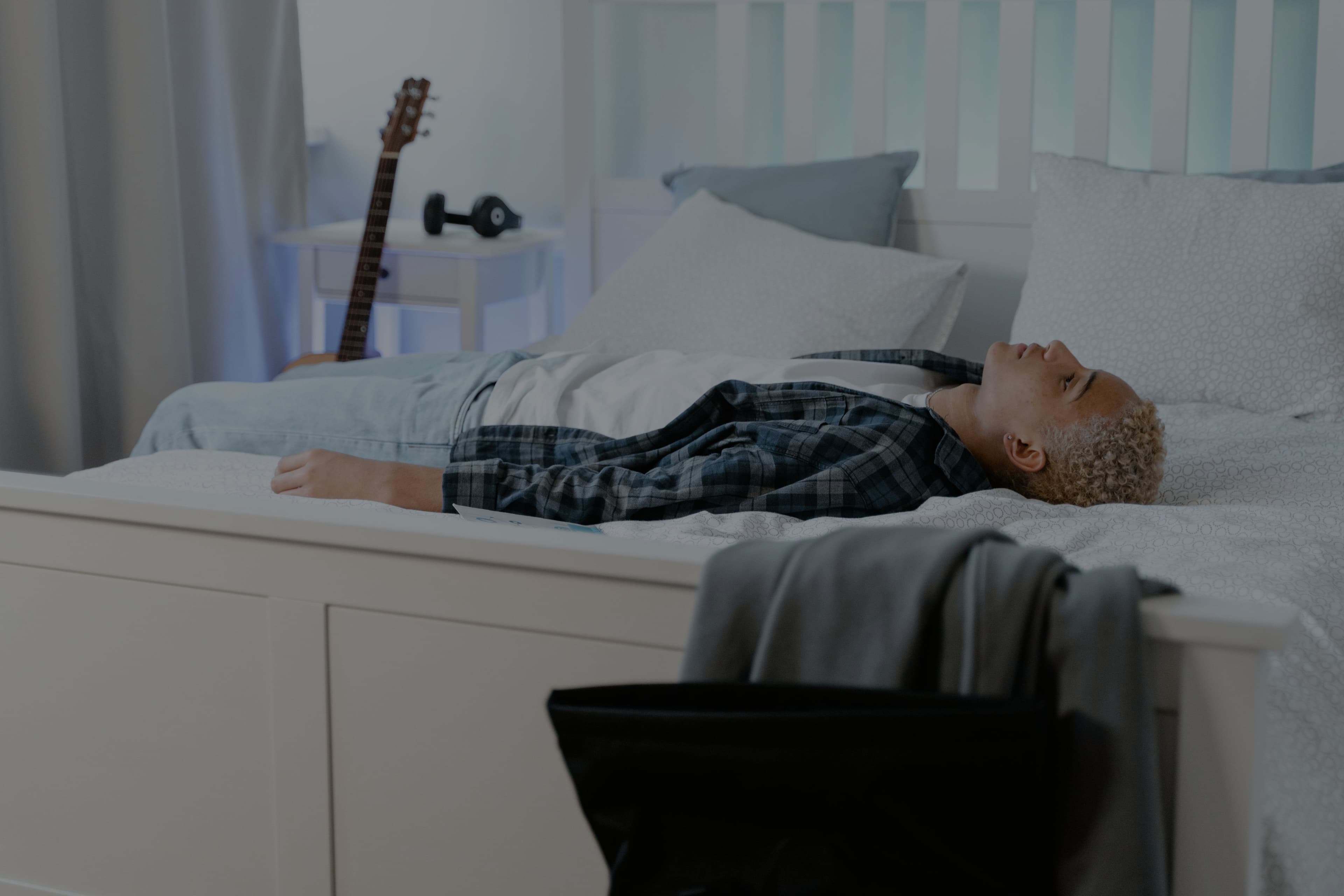A teenager lying on his back on a bed, staring up at the ceiling in a dim, quiet bedroom.