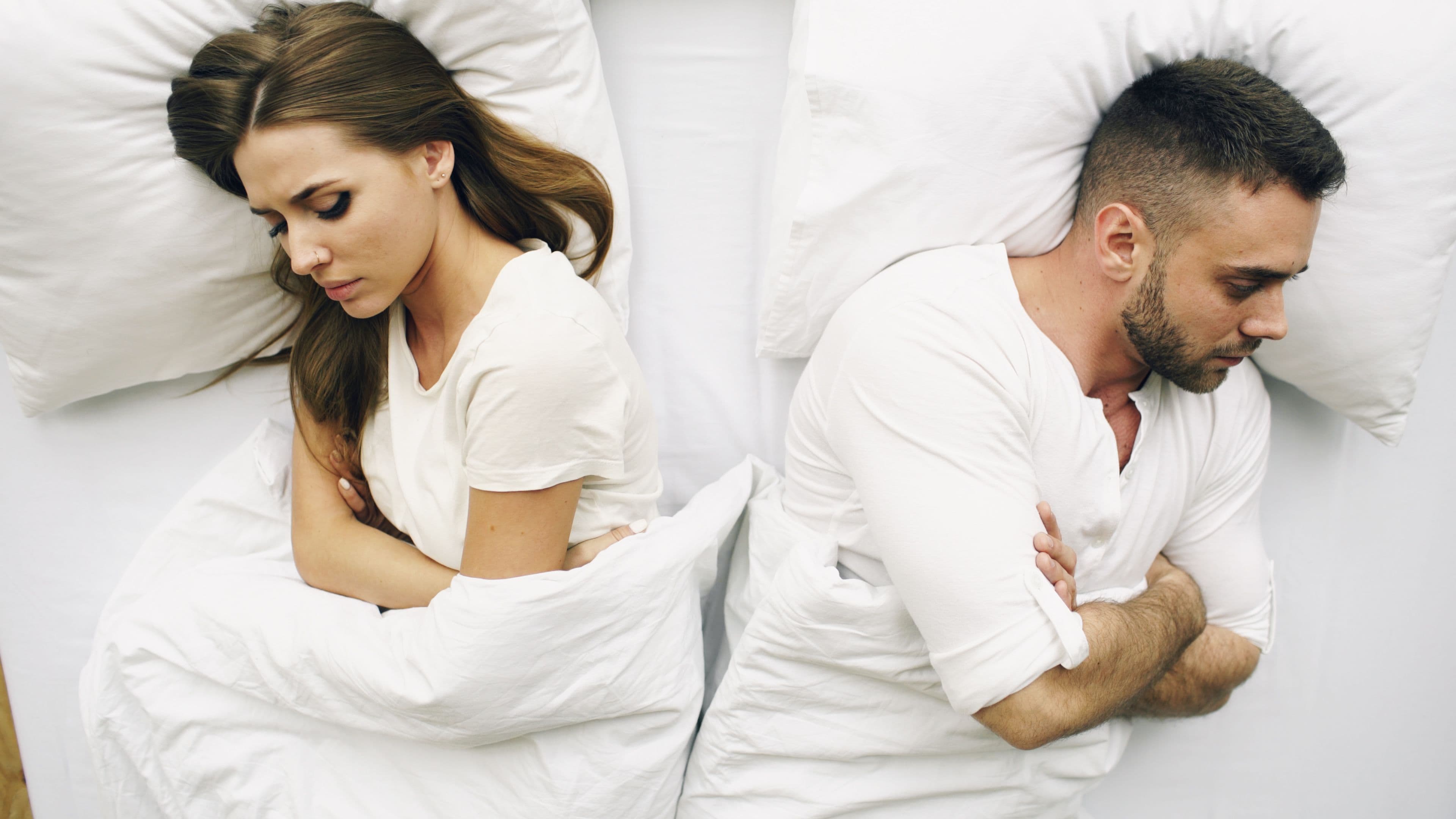 A couple lying in bed back to back, arms crossed, each looking away with an unhappy expression.