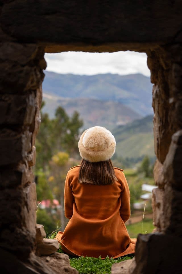 Woman in an orange coat gazing at mountains through a stone window.
